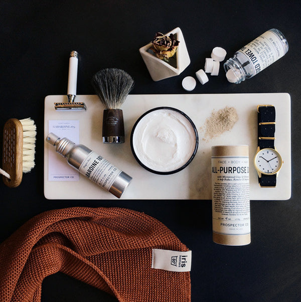 A tabletop arrangement of products including a shaving brush, razor, nail brush, watch, and towel tablets.